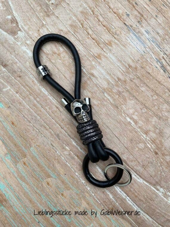 Schlüsselband mit Skull und Karabiner-Ring. 14 cm Nappaleder in Schwarz. Bestückt mit einem Ring-Karabiner in Schwarz. Dekoriert mit hochwertigem Totenkopf, Schlüsselring, Logo-Perle und Edelstahl-Endkappen. Gesamtlänge 16 cm.